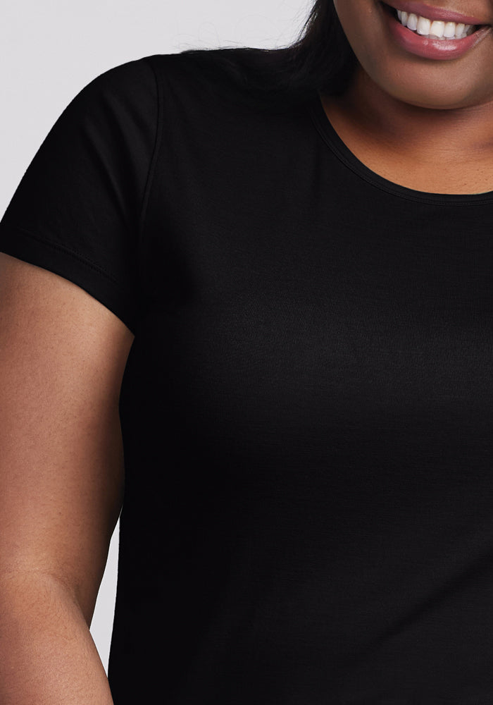 A person in a Woolx Addie Short Sleeve Crew - Black is visible from the shoulders to just above the waist. With long, dark hair and a smile, they stand against a plain light gray background, highlighting the tee's odor-free promise and casual elegance.