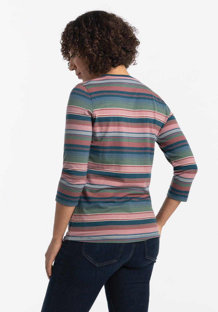 A person with curly hair is wearing a Woolx Elena V Neck - Adirondack 2.0 Stripe in shades of pink, green, and blue, paired with relaxed fit dark jeans. They are standing with their back turned, looking to the side against a plain white background.