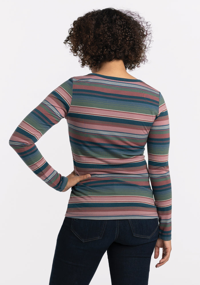 A person with curly hair is wearing the colorful Layla V Neck Top in Adirondack 2.0 Stripe from Woolx, paired with dark jeans, and stands with their back to the camera against a white background.