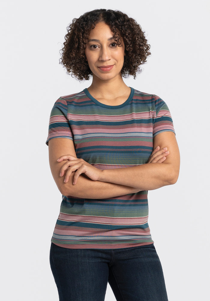 A person with curly hair stands confidently with arms crossed against a plain background, wearing the Liza Crew Neck Tee - Adirondack 2.0 Stripe by Woolx and jeans. 