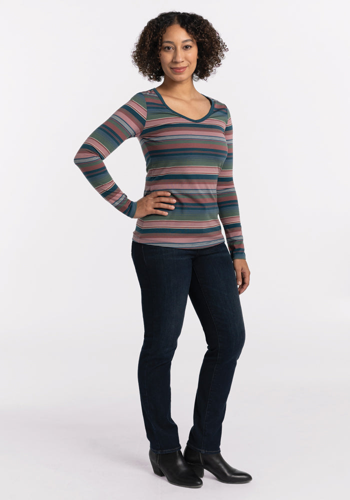 A person with curly hair is dressed in a long-sleeve, multicolored Layla V Neck Top - Adirondack 2.0 Stripe by Woolx and dark jeans. They stand against a plain background with one hand on their hip, wearing black shoes.