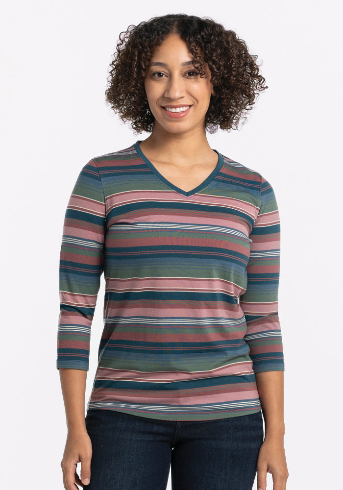 A person with curly hair smiles warmly, wearing the Woolx Elena V Neck in Adirondack 2.0 Stripe, crafted from lightweight fabric in shades of pink, green, and blue. Paired with dark jeans, they stand against a plain, light background. 