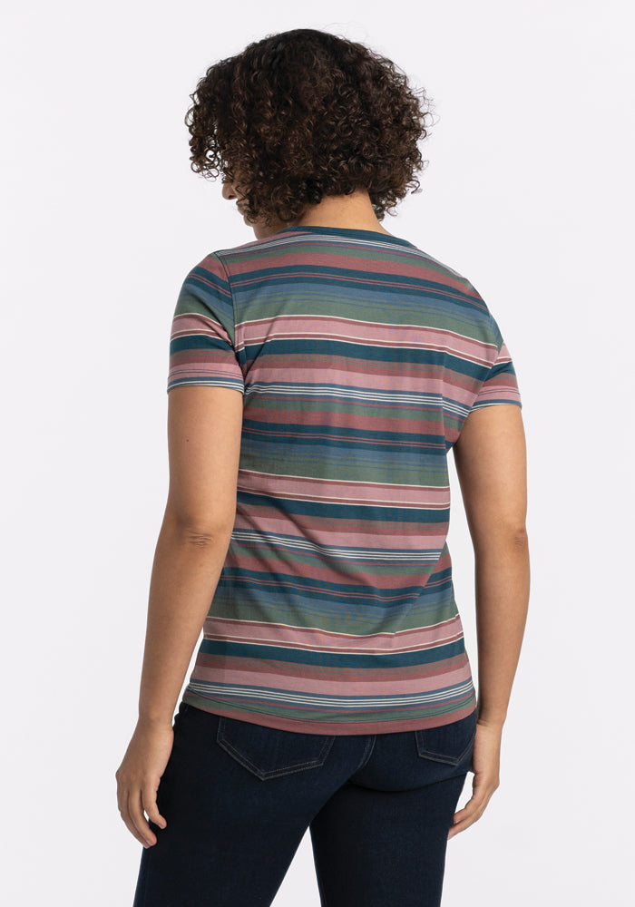 A person with curly hair stands facing away from the camera, showcasing the Woolx Liza Crew Neck Tee in Adirondack 2.0 Stripe. This lightweight merino wool tee features vibrant stripes in blue, green, pink, and brown and is stylishly paired with dark jeans against a plain white background for both style and performance benefits.