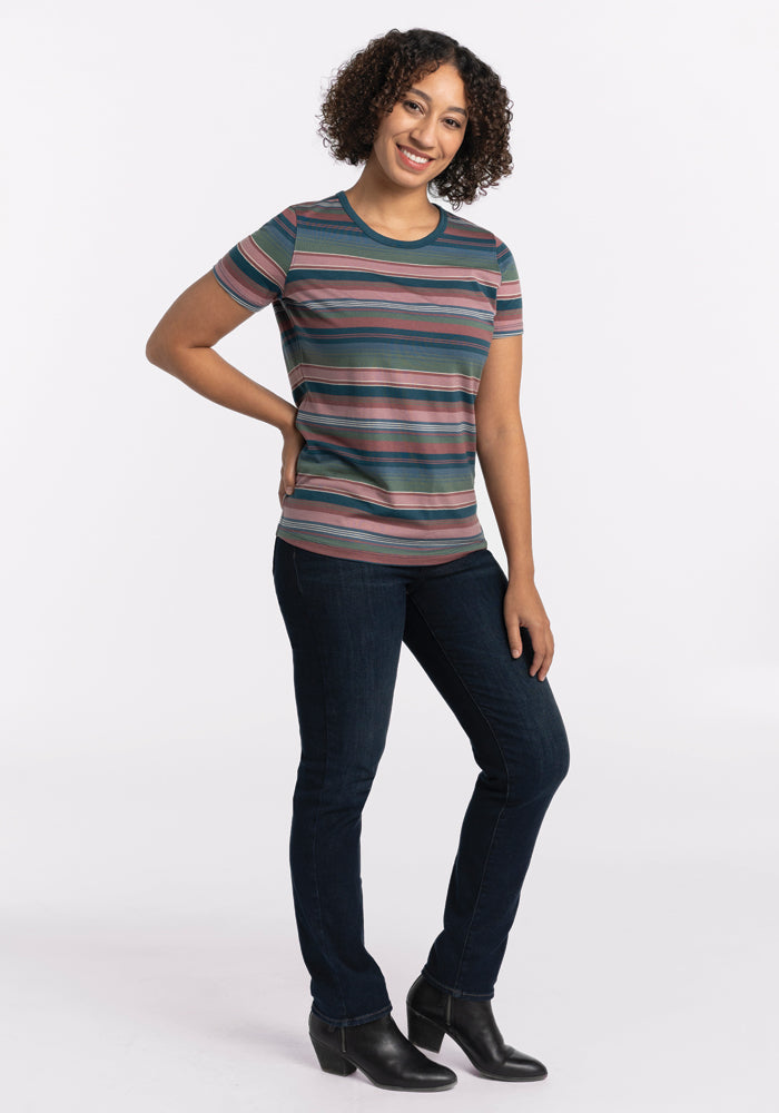 A person with curly hair smiles confidently, standing with one hand on their hip in a stylish Liza Crew Neck Tee - Adirondack 2.0 Stripe by Woolx. Paired with dark jeans and black shoes, the ensemble radiates effortless charm against a plain white background.