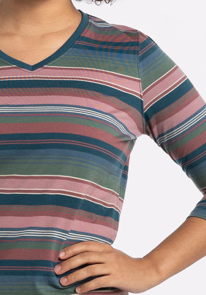 A person is wearing the Elena V Neck - Adirondack 2.0 Stripe by Woolx, featuring green, pink, blue, and white horizontal stripes. Their hand on their hip highlights the three-quarter sleeves and lightweight fabric against a plain, light-colored background that accentuates this stylish choice.