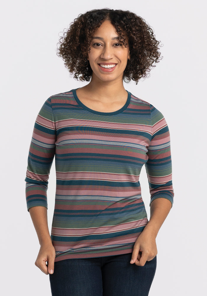 A person with curly hair smiles, wearing the Woolx Jenny 3/4 Sleeve Crew Neck in Adirondack 2.0 Stripe with dark jeans, standing against a plain, light background. 