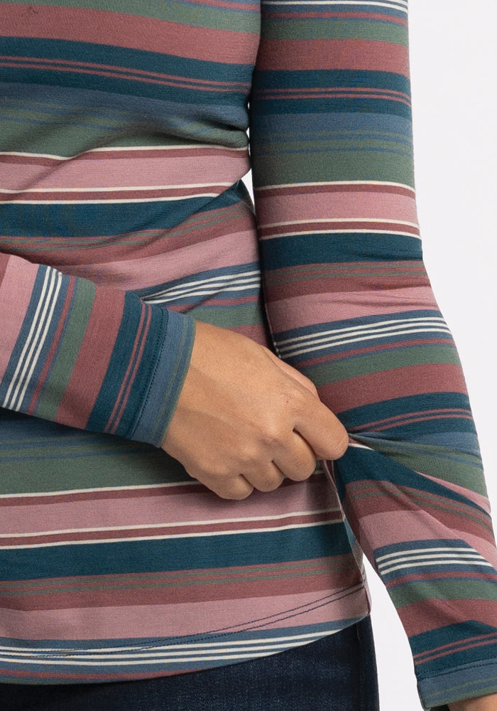 A person is wearing the Layla V Neck Top - Adirondack 2.0 Stripe by Woolx, a vibrant long-sleeved shirt crafted from merino wool, showcasing colorful horizontal stripes in shades of green, pink, blue, and maroon. The person is gently adjusting the cuff of the sleeve.