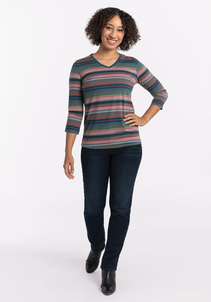 A person with curly hair is wearing an Elena V Neck - Adirondack 2.0 Stripe by Woolx, expertly crafted in lightweight merino wool, and paired with dark jeans, smiling while standing against a plain white background.