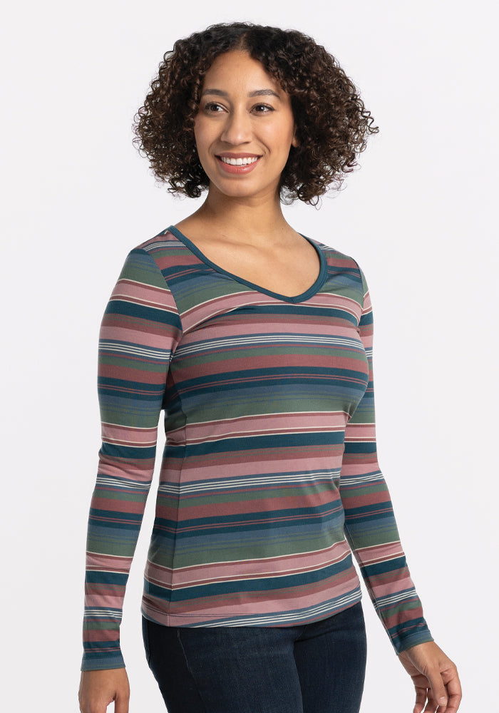 A person with curly hair is wearing the Layla V Neck Top - Adirondack 2.0 Stripe by Woolx, a long-sleeve shirt in shades of pink, blue, and green made from lightweight Merino wool. They are smiling and standing against a plain white background.