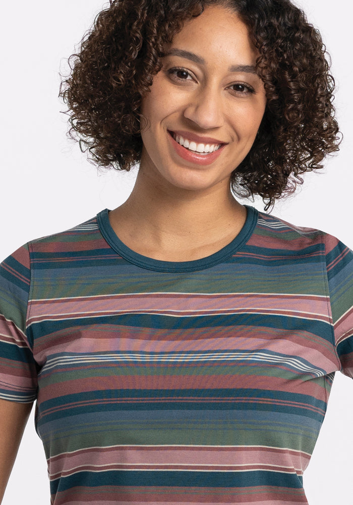 A person with curly hair smiles while wearing the Liza Crew Neck Tee - Adirondack 2.0 Stripe by Woolx, featuring a lightweight design in shades of green, pink, and blue. The merino wool fabric provides performance benefits for optimal comfort, set against a plain white background.