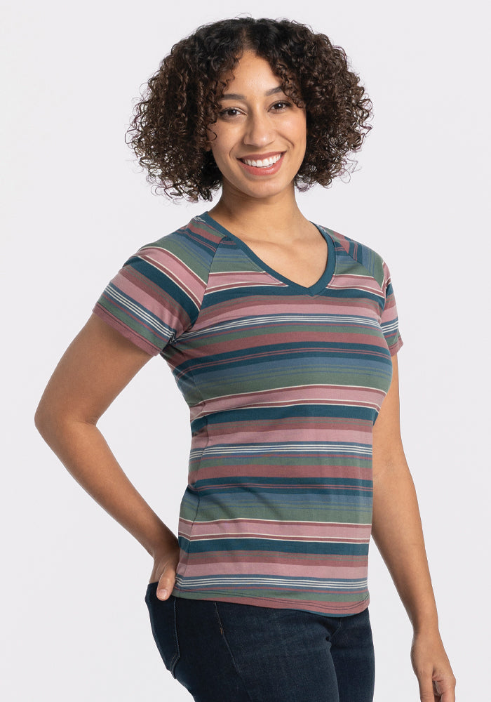 A person with curly hair smiles while wearing the vibrant, short-sleeved Mia V Neck - Adirondack 2.0 Stripe shirt by Woolx, paired with jeans. They are standing against a plain white background. 