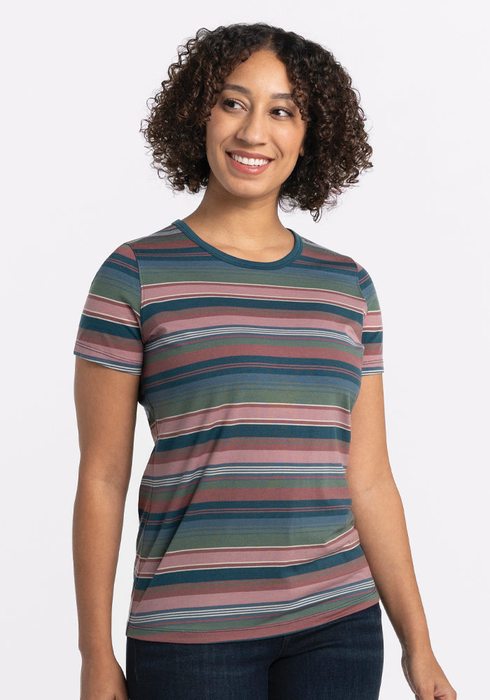 A person with curly hair smiles while wearing a short-sleeved Liza Crew Neck Tee in Adirondack 2.0 Stripe by Woolx, paired with dark jeans. The background is plain white.