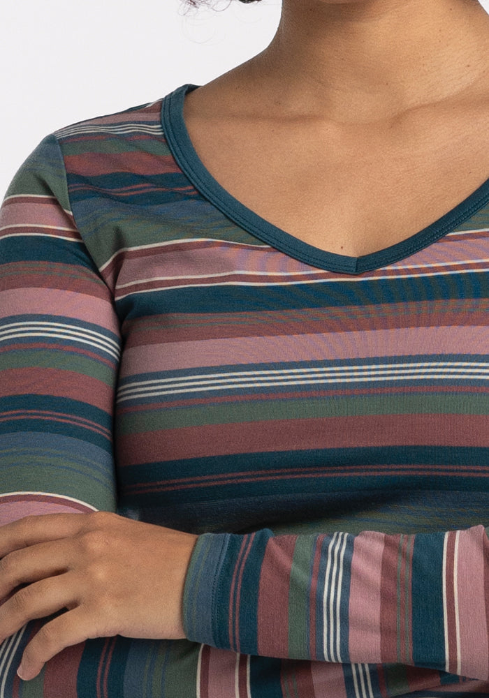 A person with curly hair is wearing the Woolx Layla V Neck Top in the Adirondack 2.0 Stripe design. This long-sleeved performance tee features horizontal stripes in green, blue, pink, and burgundy. Their arms are crossed, and the image is cropped just below the shoulders.