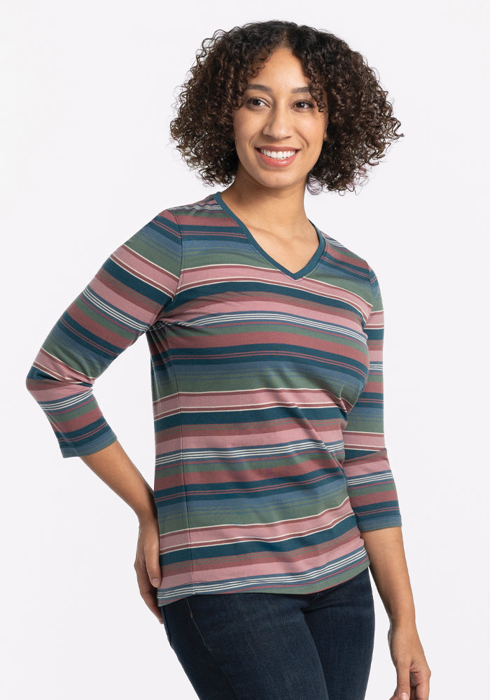 A person with curly hair is smiling and wearing the relaxed fit Elena V Neck - Adirondack 2.0 Stripe top from Woolx, featuring shades of pink, blue, and green. The striped 3/4 sleeve shirt is made from soft Merino wool as they stand against a plain white background with one hand on their hip.