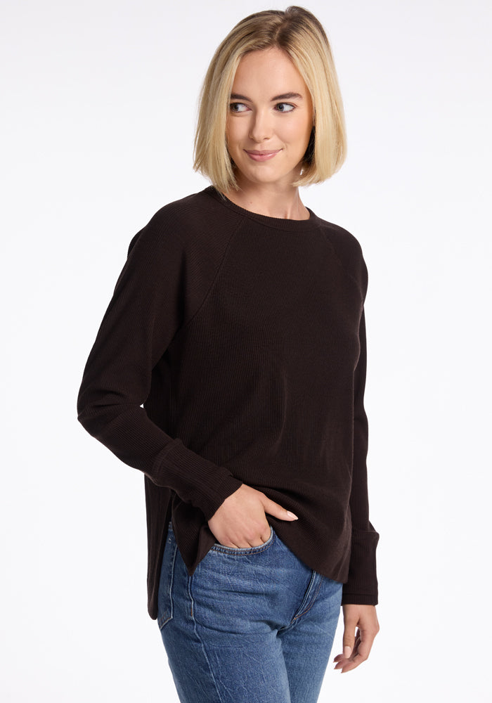 A person with shoulder-length blonde hair looks to the side while smiling, wearing a Woolx Ainsley Sweater and blue jeans with one hand resting in their pocket. Their dark long-sleeve top complements a versatile style, set against a plain white background.