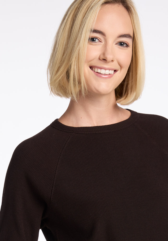 A person with shoulder-length blonde hair smiles at the camera, wearing a dark, long-sleeved Ainsley Sweater by Woolx made of Merino wool. The plain, light background highlights the individual's versatile style beautifully.