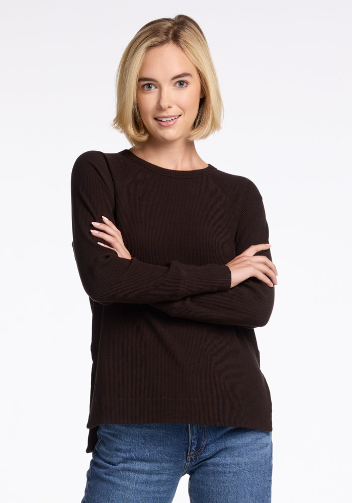 A person with short blonde hair is standing with arms crossed, smiling at the camera. They are wearing a versatile Ainsley Sweater made of black Merino wool from Woolx, paired with blue jeans. The background is white. 