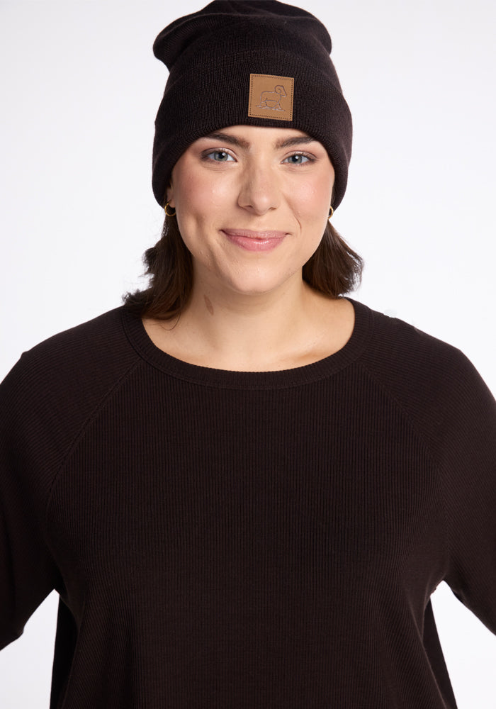 A person with shoulder-length dark hair is smiling while wearing a black beanie with a small patch and the versatile Ainsley Sweater by Woolx. The background is plain and well-lit, emphasizing the person's face and clothing.