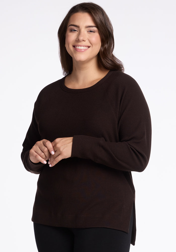 A person with shoulder-length brown hair smiles while standing in front of a plain background. They are wearing a long-sleeved, dark Ainsley Sweater - French Roast from Woolx made of Merino Wool and holding their hands together at chest height, showcasing a versatile style. 