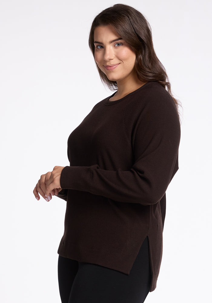 A woman with long, wavy dark hair is smiling while wearing a dark brown, long-sleeved Merino wool sweater by Woolx. She is posing with her body slightly angled to the side and her hands softly clasped in front. The versatile style of the Ainsley Sweater stands out against the plain white background.