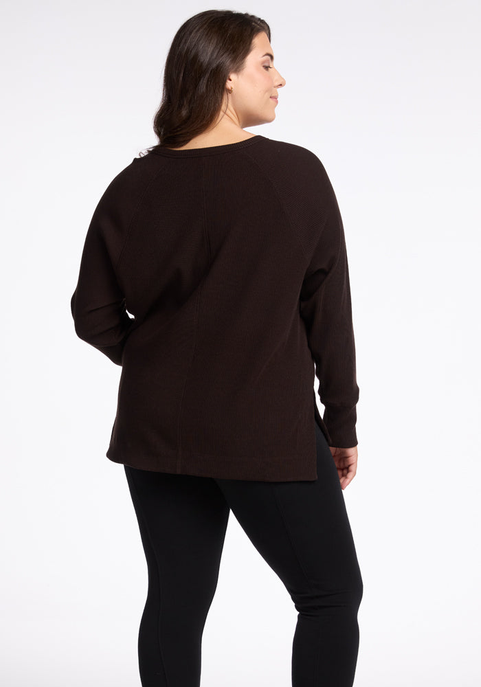 A person with long hair is shown from the back, wearing a dark, long-sleeved Ainsley Sweater by Woolx and black pants. They are standing against a plain white background, epitomizing versatile style.