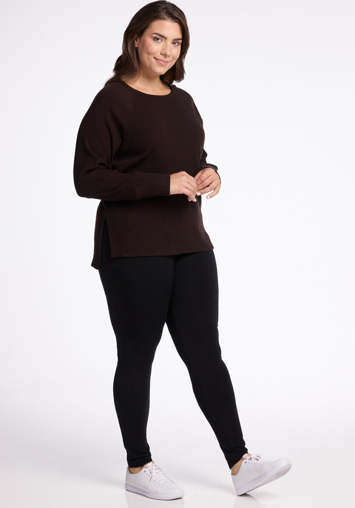 A person with medium-length brown hair stands against a white background, smiling slightly. They are wearing a long-sleeved Ainsley Sweater in dark brown from Woolx, made of Merino Wool, paired with black leggings and white sneakers. Their hands are gently clasped in front, showcasing their versatile style.