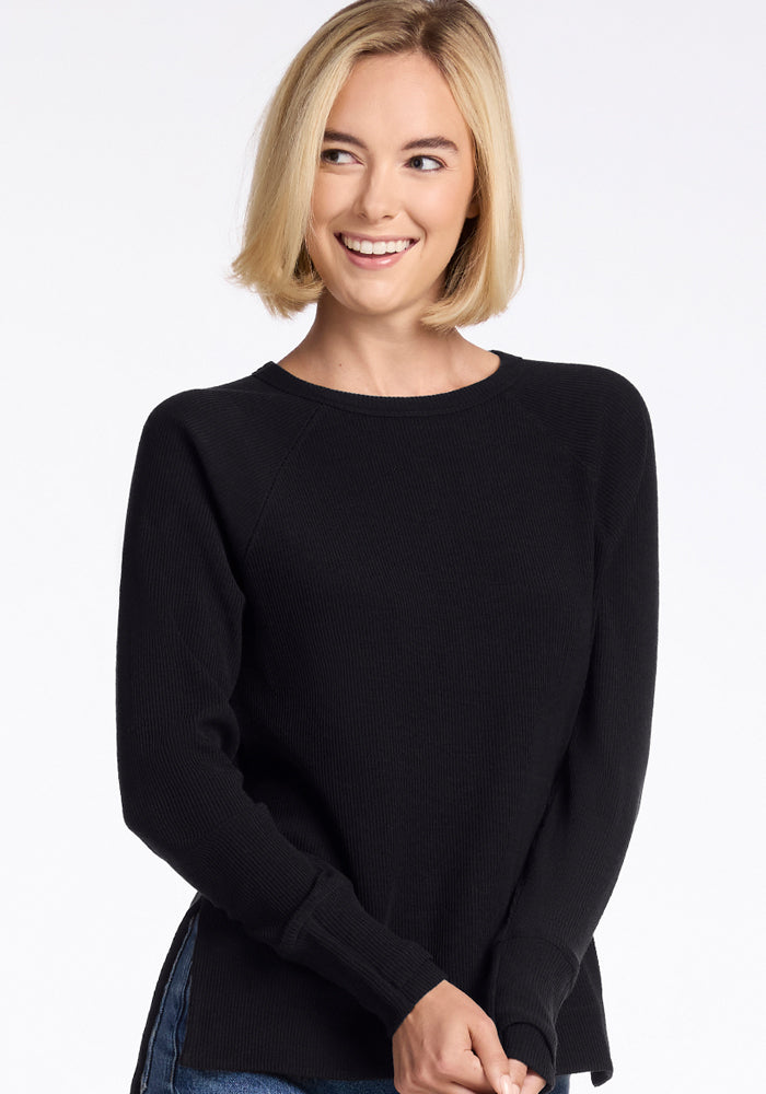 A person with short, blonde hair is smiling while wearing the Woolx Ainsley Sweater in black paired with blue jeans. The background is plain white.  