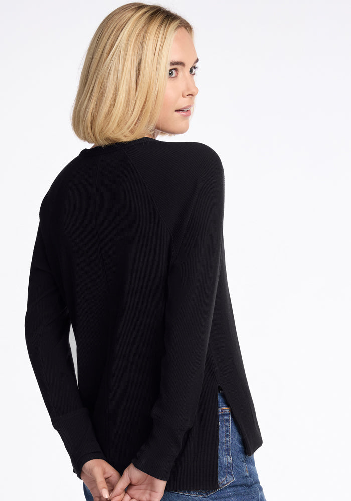 A woman with shoulder-length blonde hair, dressed in a temperature-regulating Woolx Ainsley Sweater in black and blue jeans, stands facing away but gazes over her right shoulder. Her hands are positioned behind her back against a plain white canvas backdrop.