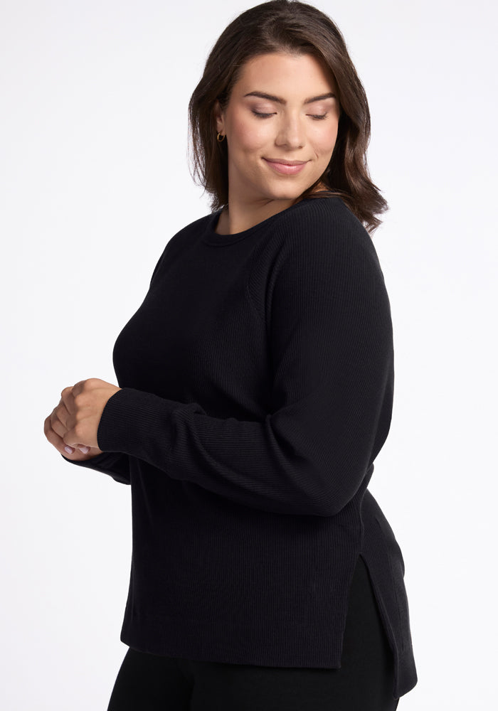 A woman with long brown hair is smiling with her eyes closed. She is wearing the Ainsley Sweater in black from Woolx, which features ribbed detailing for added texture, and stands against a plain white background.