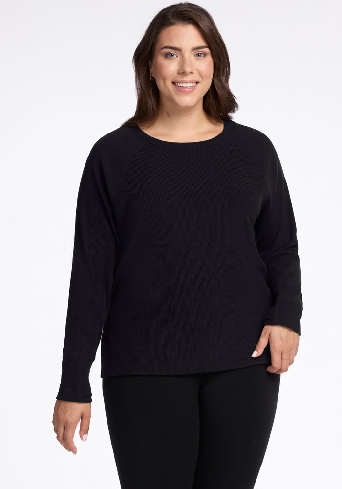 A woman with long brown hair smiles, wearing the Woolx Ainsley Sweater in black, crafted from temperature-regulating Merino wool, along with matching pants, as she stands against a plain white background. 