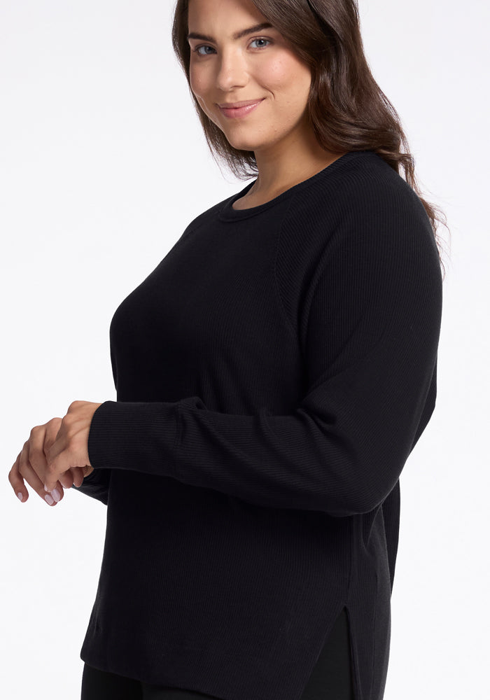 A woman with long dark hair is wearing the Ainsley Sweater in black by Woolx, featuring ribbed detailing. She is smiling softly and standing against a plain white background.