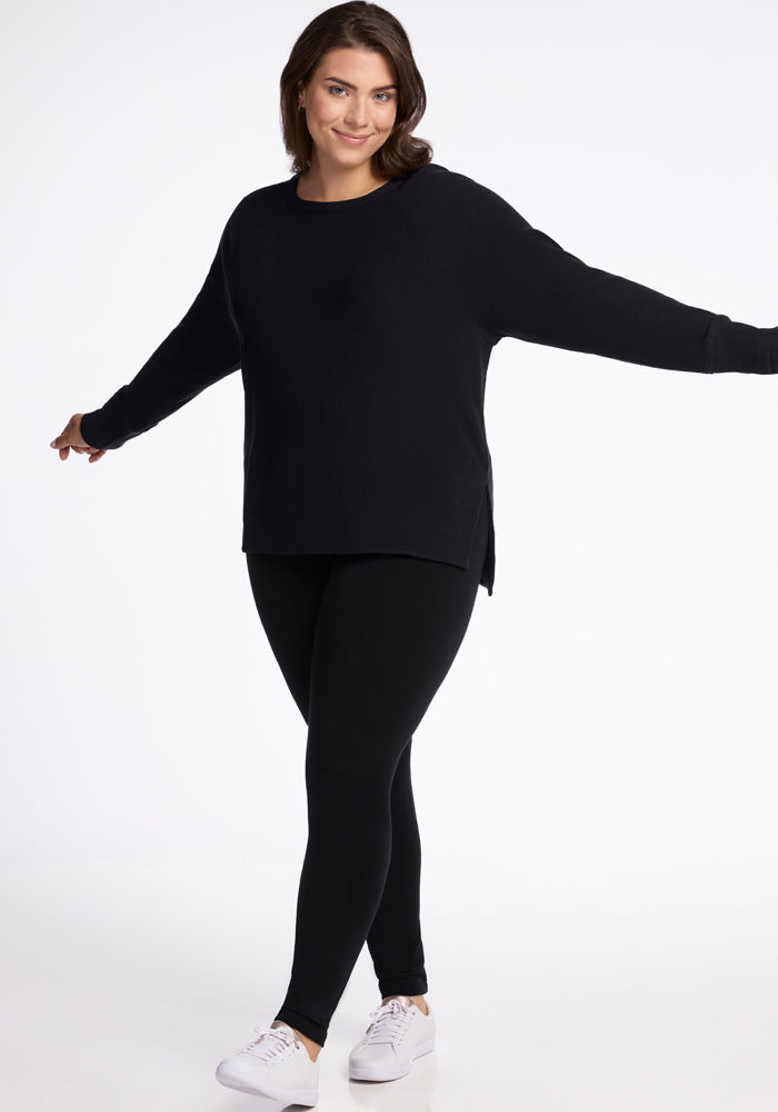 A person wearing the Ainsley Sweater in black from Woolx, paired with black leggings, smiles with arms slightly outstretched. They are sporting white sneakers and stand against a plain white background.