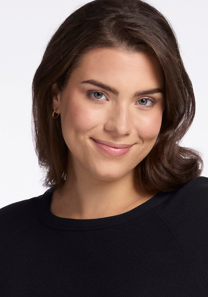 A person with shoulder-length brown hair smiles softly, adorned in the Ainsley Sweater - Black by Woolx, featuring elegant ribbed detailing. The plain white background offers a clean and simple setting that accentuates the understated sophistication of this temperature-regulating Merino wool piece.