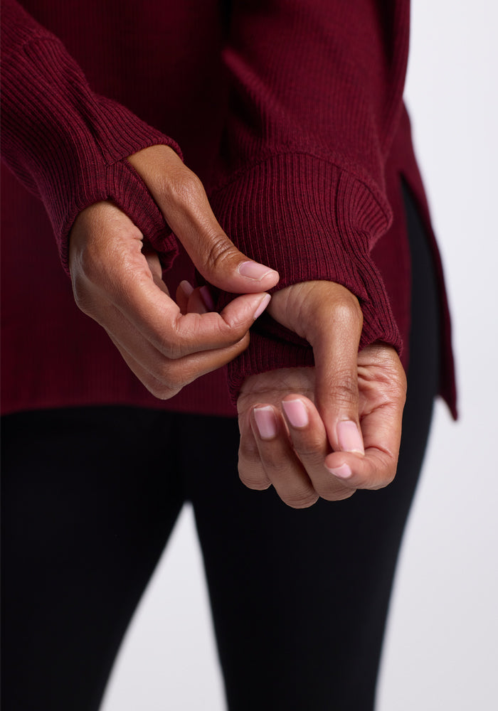 A person is shown from the waist down, wearing a burgundy long-sleeve Ainsley Sweater with thumb holes in the sleeves. They are pulling on the sleeve with both hands. The sweater, made of soft Merino wool for a versatile style, is from Woolx and is paired with black pants against a plain, light-colored background.