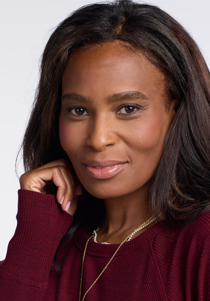 A woman with long, dark hair is smiling softly at the camera. She is wearing a maroon Ainsley Sweater by Woolx and a gold necklace. Her right hand gently rests against her face. The background is plain and light-colored, highlighting her versatile style in the elegant Ainsley Sweater.