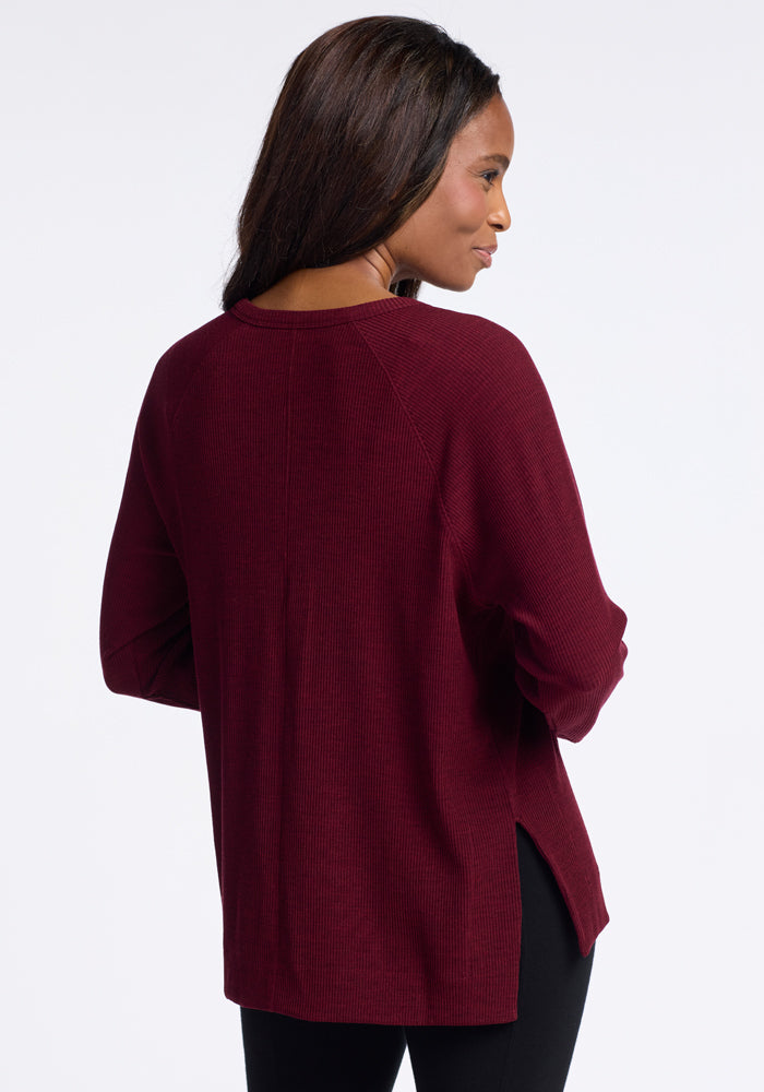 A woman with long dark hair is slightly turned to her right, showcasing the back of a long-sleeved, burgundy Ainsley Sweater by Woolx. She is also wearing black pants against a plain white background.