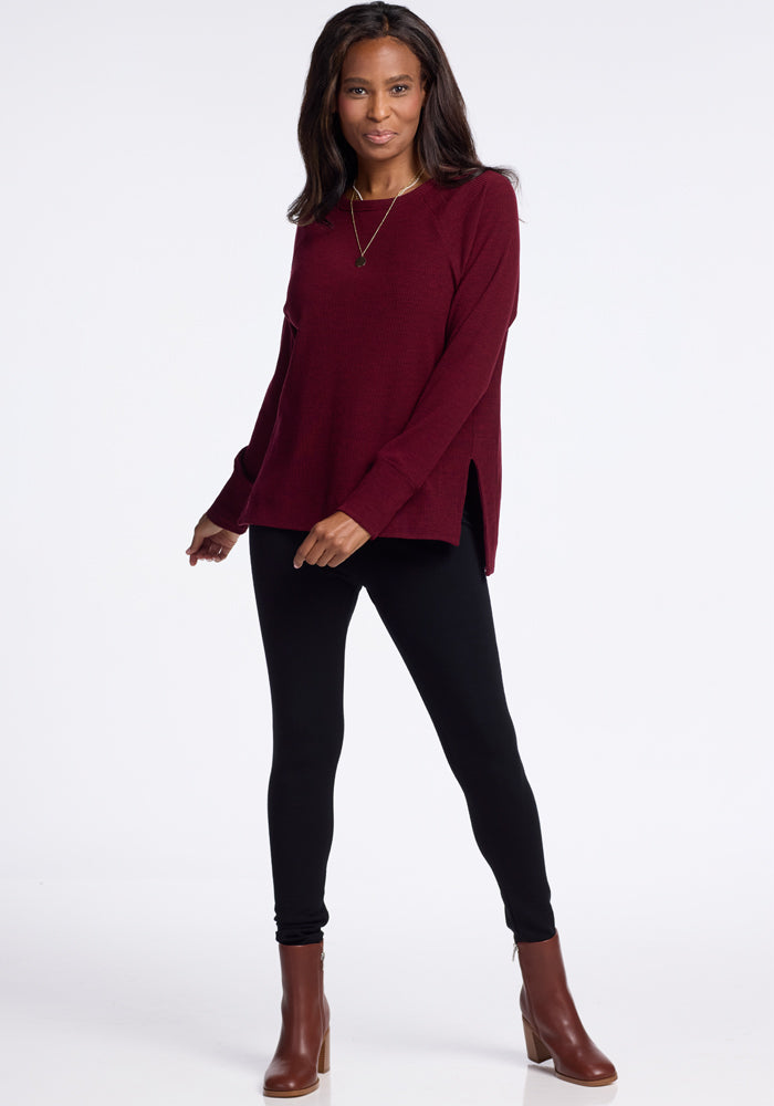 Woman stands against a plain white background, wearing a long-sleeved burgundy Woolx Ainsley Sweater, black leggings, and brown heeled ankle boots. She has long dark hair and wears a necklace. She is smiling slightly and has her arms relaxed by her sides, showcasing her versatile style.