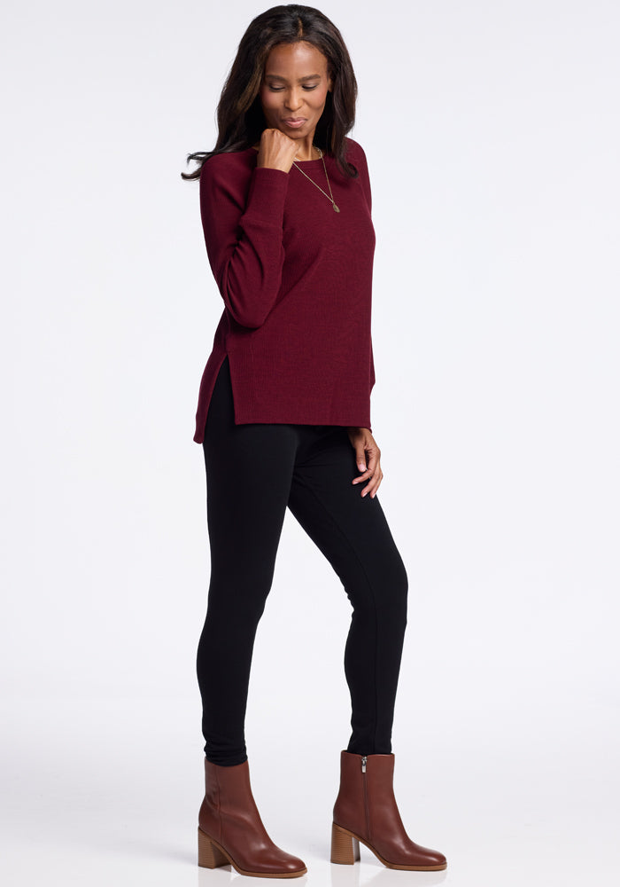 A woman stands on a plain background, wearing a maroon Ainsley Sweater by Woolx made of soft merino wool, black leggings, and brown ankle boots with a block heel. She has long, wavy hair and is looking down with one hand touching her chin. Her versatile style is completed with a pendant necklace.