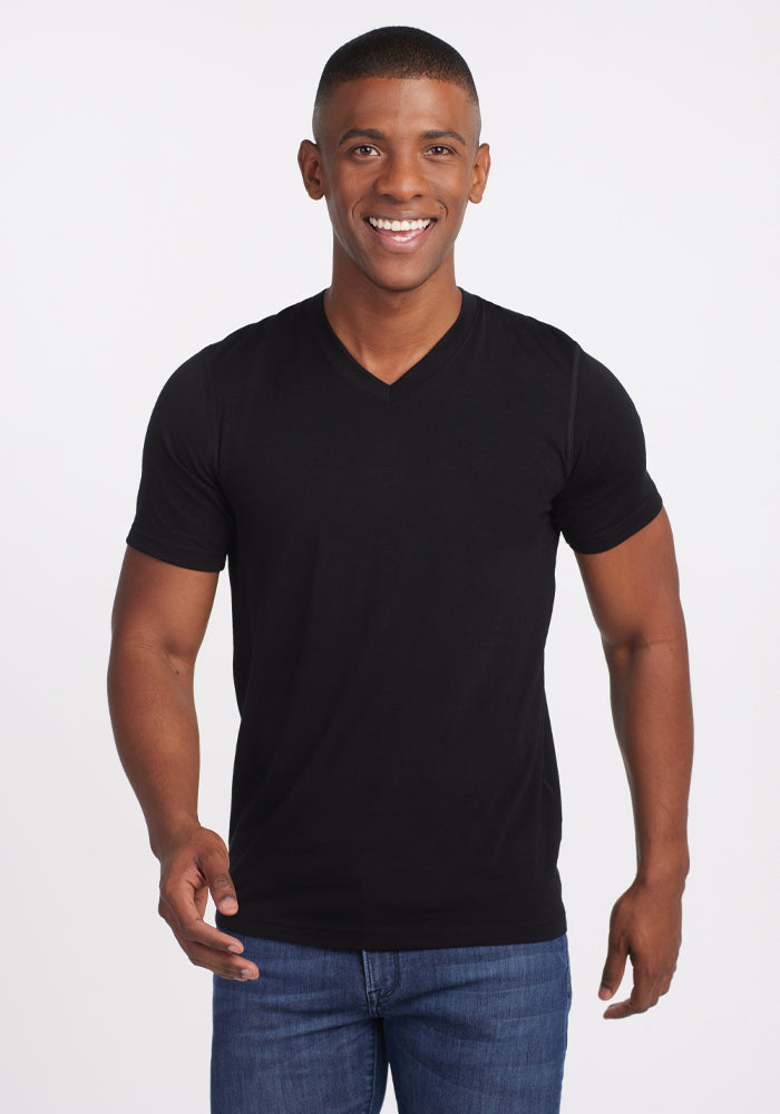 A person smiling and standing against a plain white background, wearing the Ashton V Neck in black by Woolx paired with blue jeans, enjoying the comfort of the lightweight, odor-free Merino fabric. 