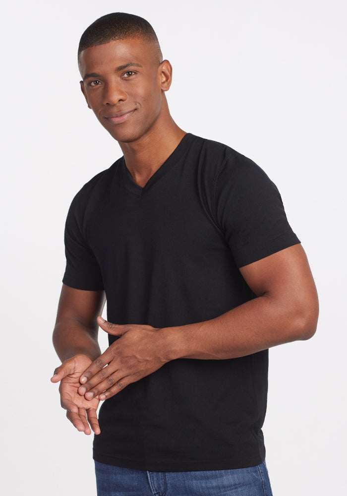 A person wearing the Ashton V Neck in black by Woolx and blue jeans poses against a plain white background. The odor-free garment complements their short hair and soft smile as they clasp their hands in front of them.