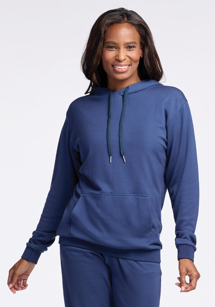 A person wearing a Woolx Avery Hoodie - Starry Night from the WoolAire Collection, made of soft merino wool, and matching blue pants is standing and smiling. The hoodie features drawstrings and a front pocket. The background is plain white. 