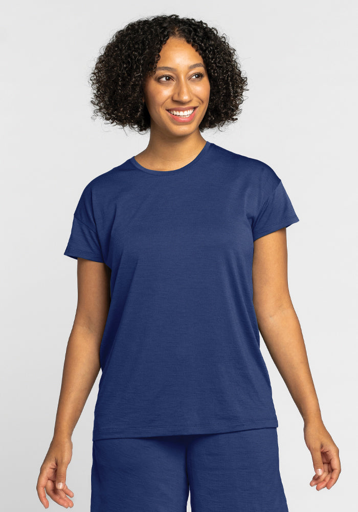 A person with curly hair smiles, comfortably clad in a Woolx Billie Lounge Tee in the Starry Night color and matching pants against a light background.