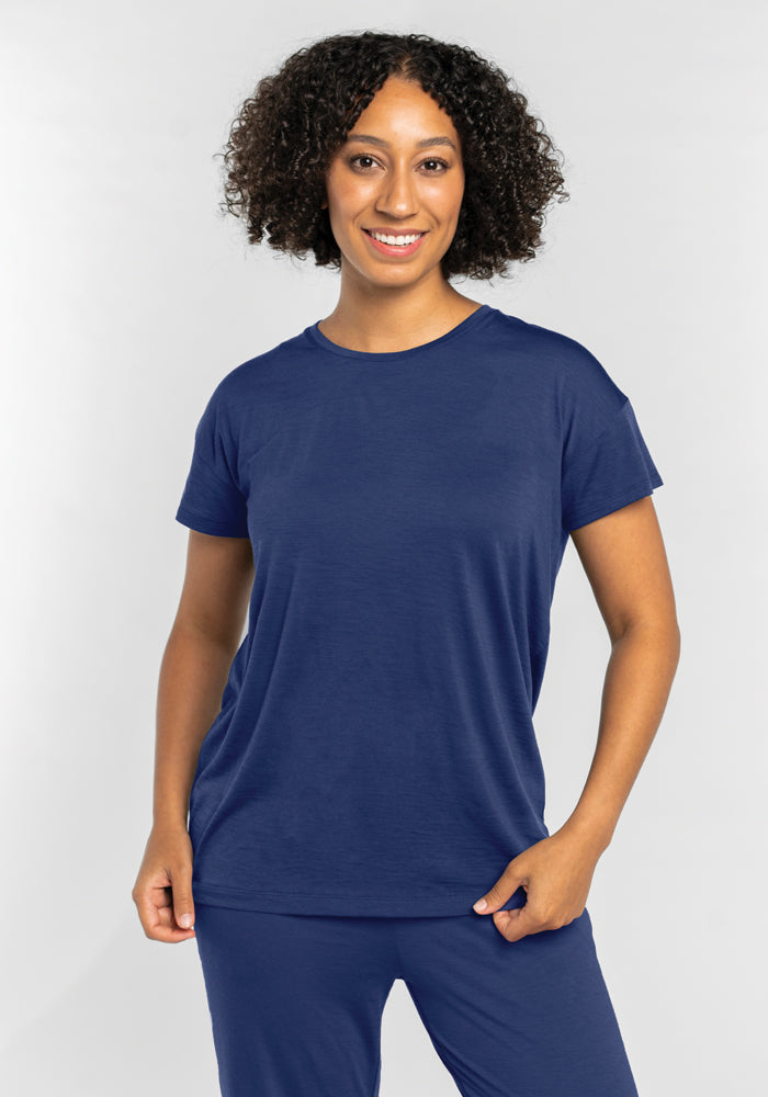 A person with curly hair smiles in the temperature-regulating and moisture-wicking Billie Lounge Tee - Starry Night by Woolx, standing against a plain light gray background. 