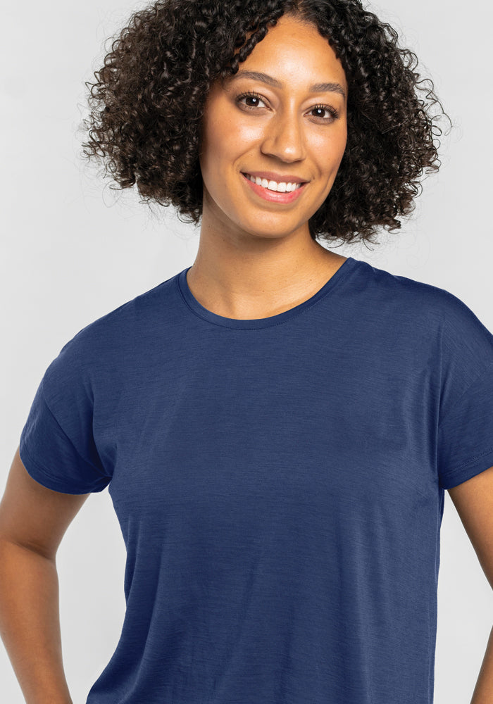 A person with curly hair smiles while wearing the Billie Lounge Tee in Starry Night from Woolx, crafted from cooling merino wool, ideal for hot sleepers. The background is a light gray.