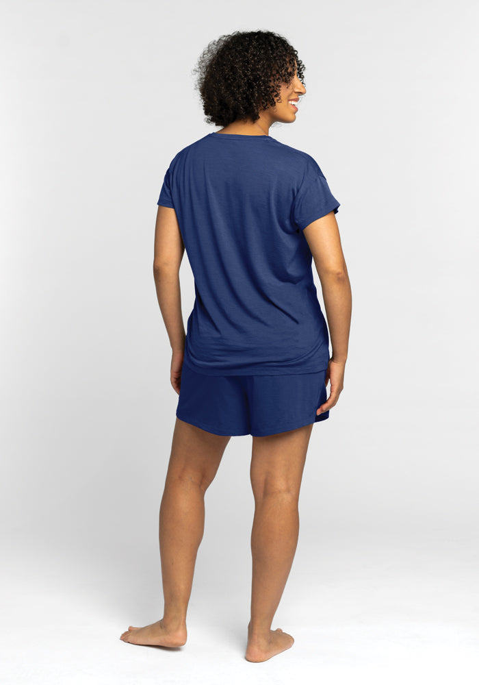 A person with curly hair is standing barefoot, facing away, wearing the Billie Lounge Tee in Starry Night and matching moisture-wicking shorts against a plain light gray background.