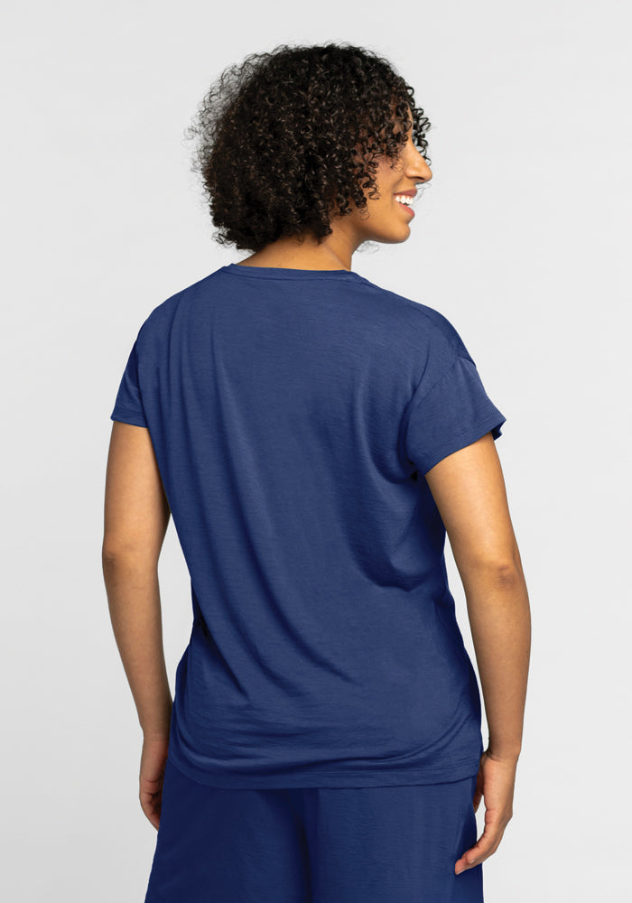 A person with curly hair is standing and facing away from the camera, wearing a Billie Lounge Tee in Starry Night by Woolx and blue pants. The background is plain and light-colored.