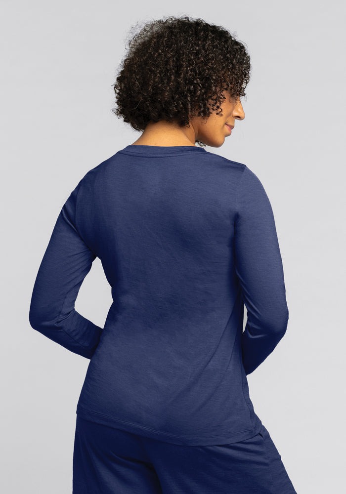 A person with curly hair is shown from behind, wearing the Woolx Lily V Neck Lounge Top in Starry Night and matching pants made from moisture-wicking fabric. The background is a plain light gray.