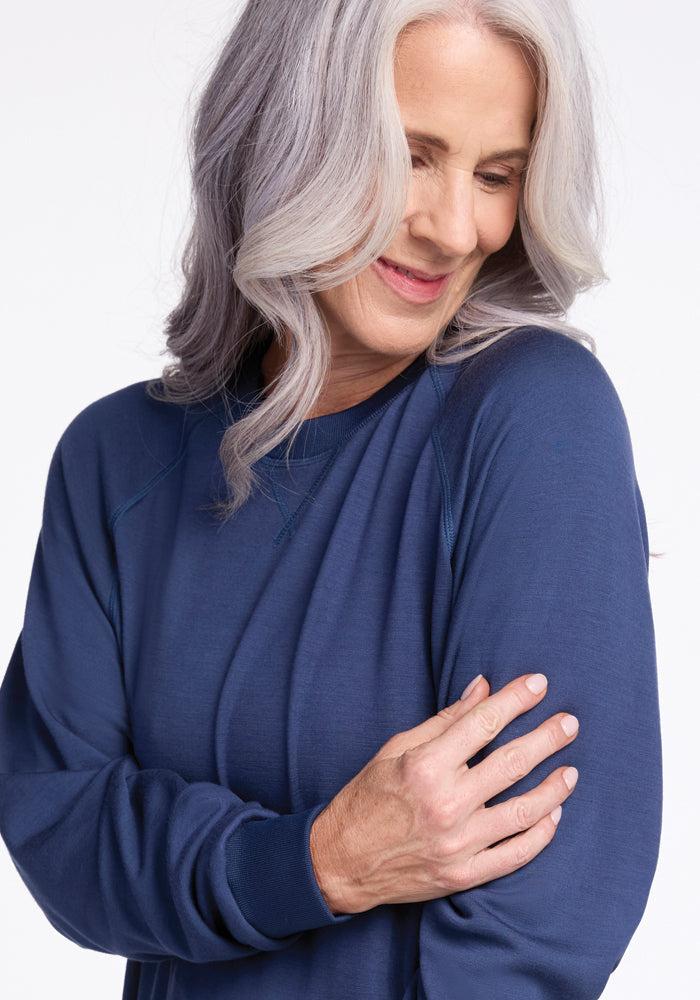 A woman with long, wavy gray hair smiles gently while looking down. She wears the Bailey Crewneck Sweatshirt in Starry Night from Woolx, crafted with soft Merino Wool. Her right arm is gently held by her left hand against a plain, light background.