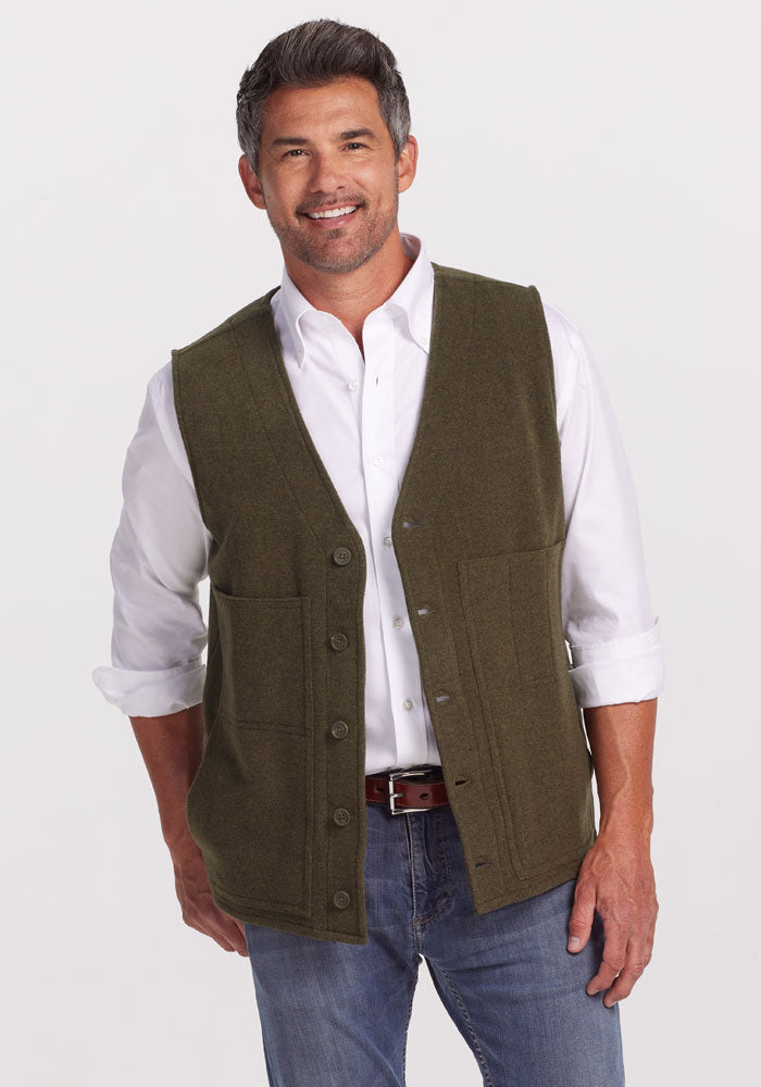 A man smiles in front of a plain white background, wearing the Woolx Baker Vest in Dark Forest over a white shirt, paired with blue jeans.