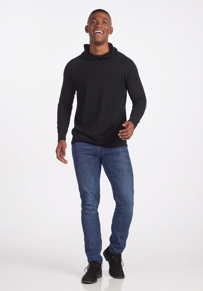 A person wearing a stylish black Base Camp Hoodie by Woolx and blue jeans stands smiling against a plain white background. They walk forward with a relaxed posture, exuding the comfort and style of outdoor apparel.